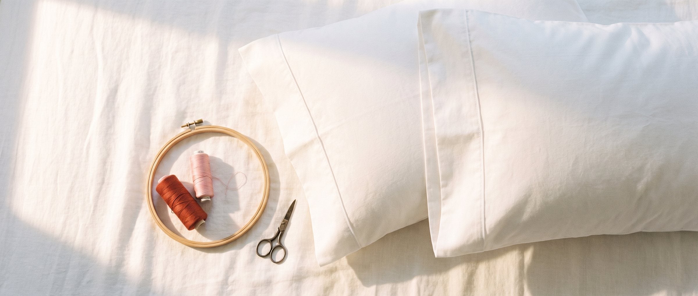 Taies d’oreiller en percale blanche à plat, tambour à broder vide et bobines de fil rouge et rose sur du lin