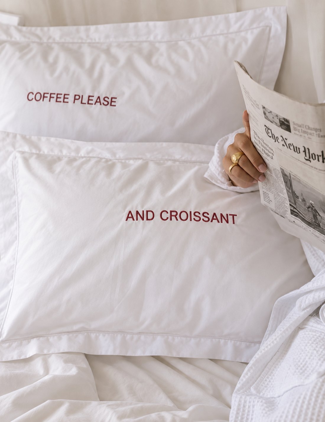 Deux taies d’oreiller brodées « COFFEE PLEASE » et « AND CROISSANT », un journal à la main
