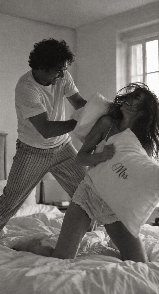 Un couple en pyjama se lance dans une bataille d’oreillers sur un lit défait, une taie brodée « Mrs. » entre eux, photo en noir et blanc