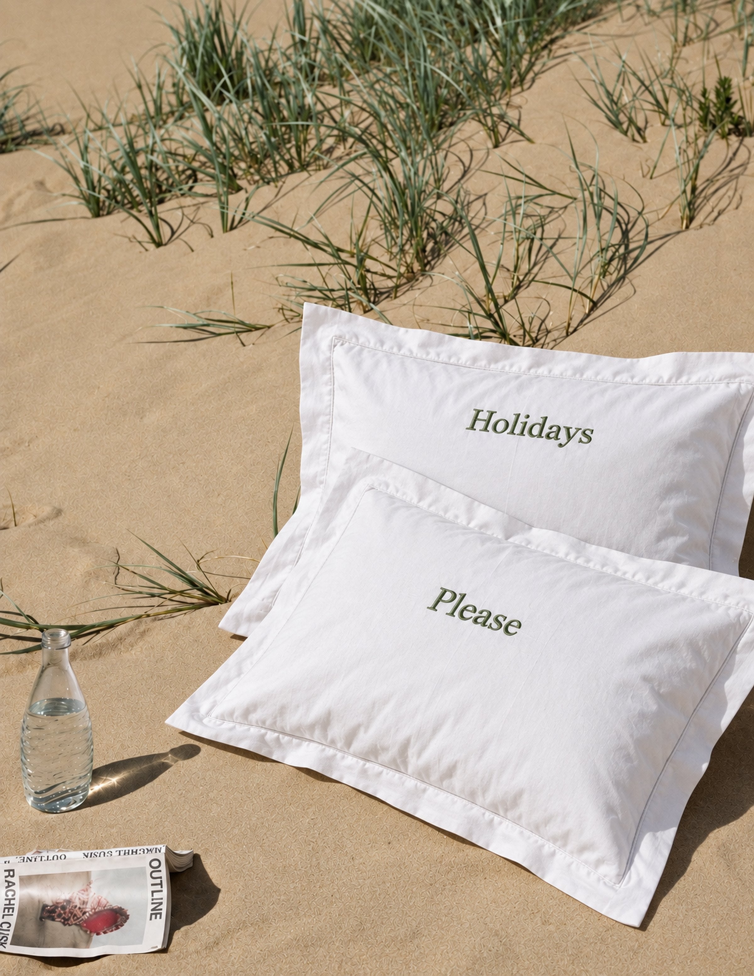 Deux taies d’oreiller blanches brodées « Holidays » et « Please », posées sur le sable