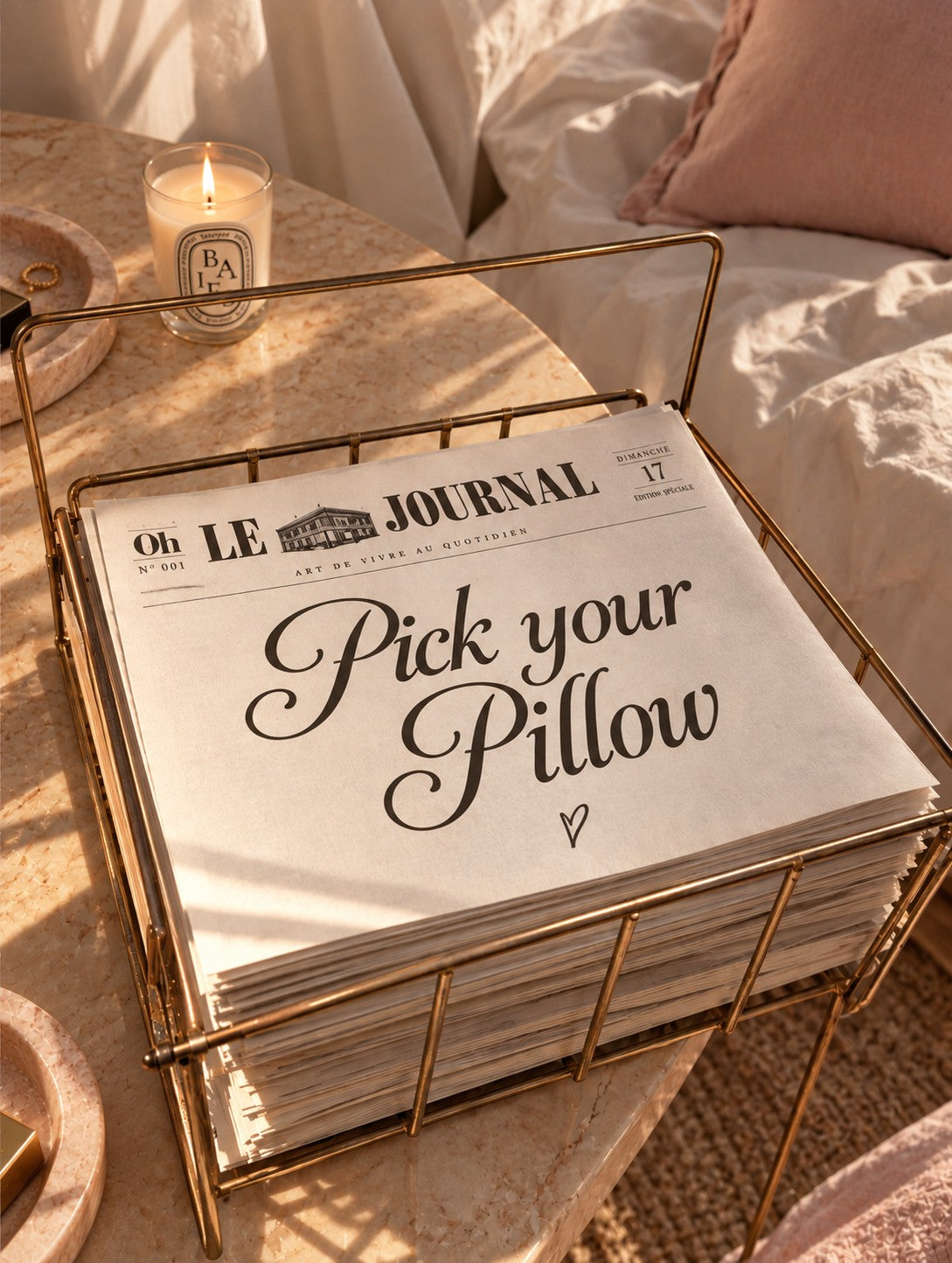 Un exemplaire du journal « Pick your Pillow » posé sur une table de chevet en marbre rose, bougie allumée, coin d’une taie d’oreiller personnalisée rose en arrière-plan